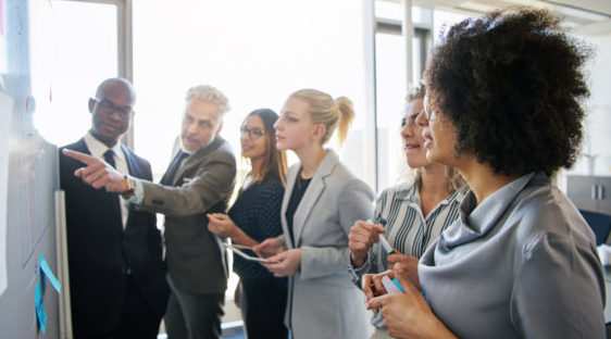 Group of business people standing around a whiteboard reviewing tactics for their annual plan.