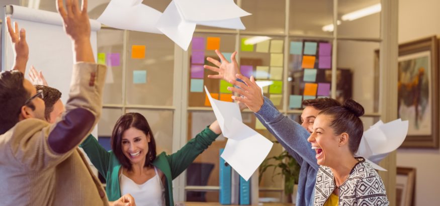 five co-workers sitting around a conference table laughing and smiling while throwing papers in the air